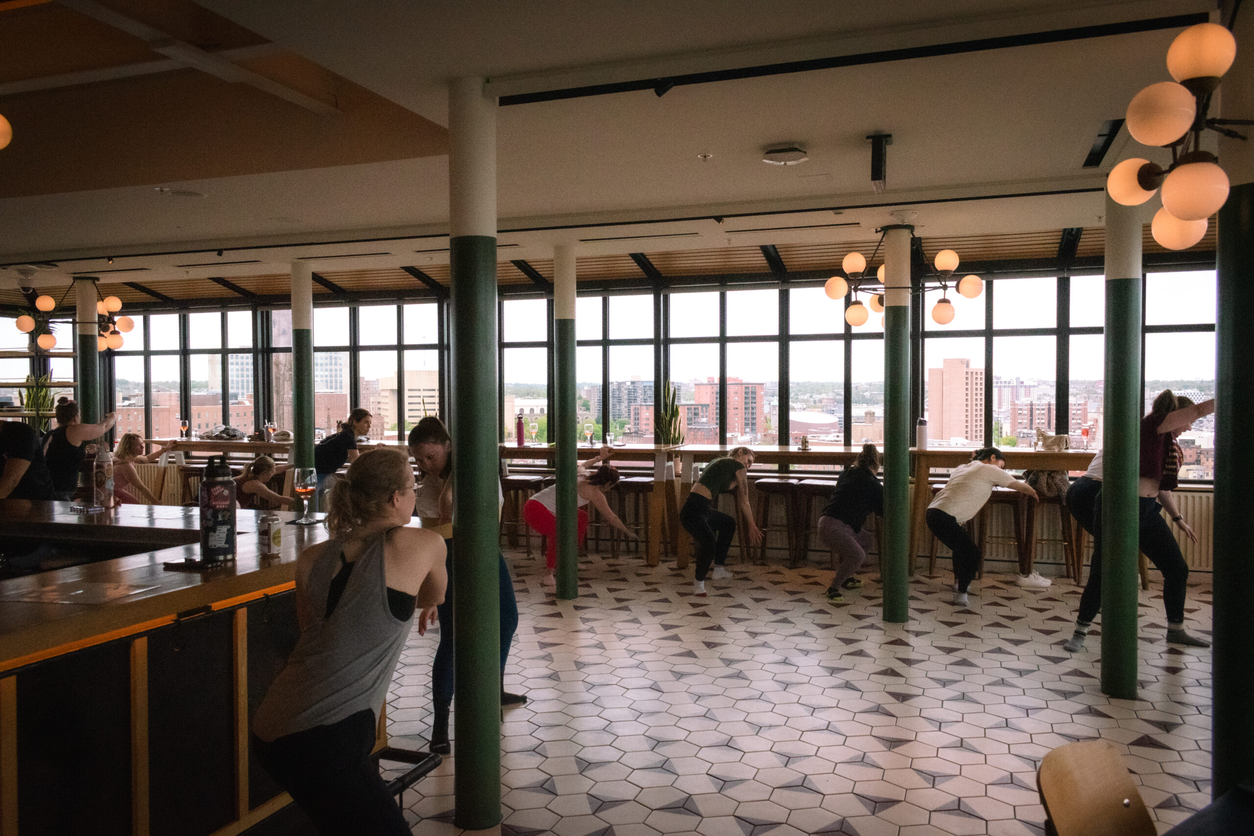 Fitness class participants exercising in modern rooftop studio with city skyline views and overhead lighting.